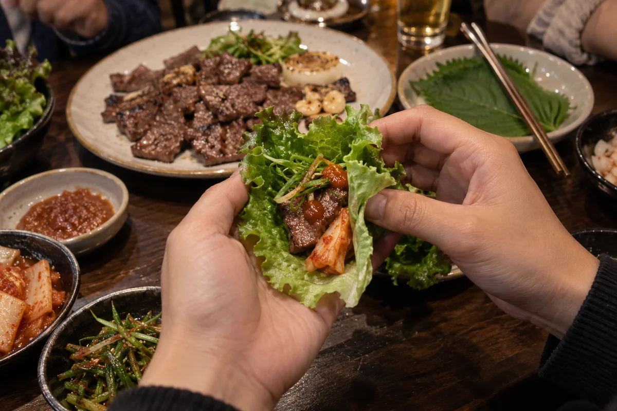 A diner gently folds a lettuce leaf around grilled beef, ssamjang, scallions and a little kimchi using both hands.